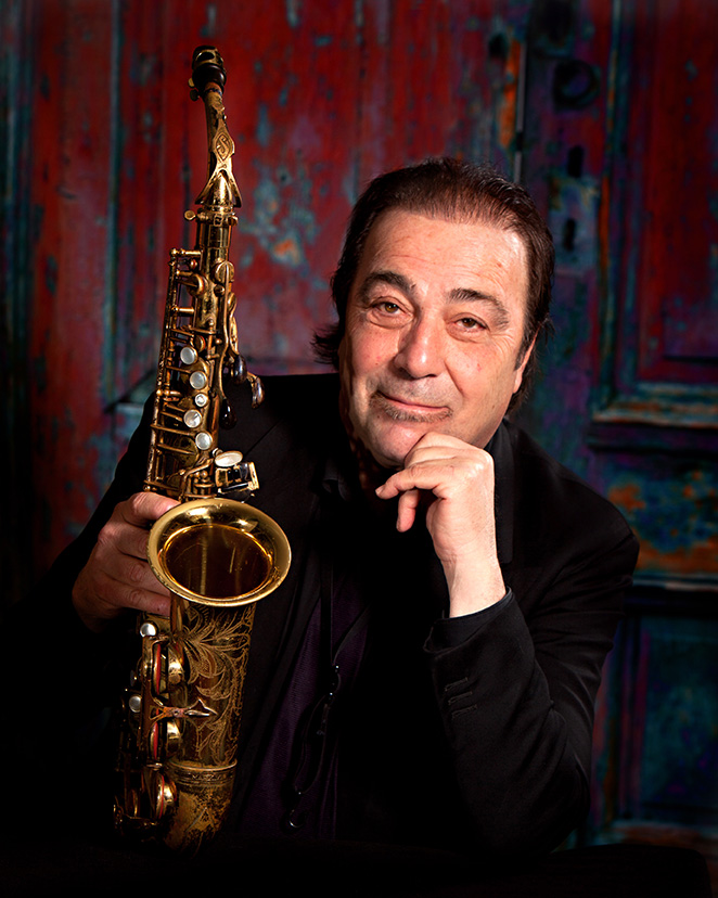 Portrait of Greg Abate from the waist up looking directly into the camera. He holds his saxphone in his left had and his right hand is tucked under his chin.