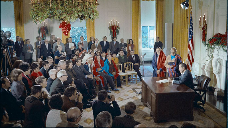Archival image of President Nixon in the Oval Office surrounded by Taos Pueblo delegates and others at the formal bill signing.