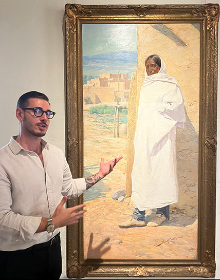 Curator Christian Weguespack gestures in front of a painting featuring a indigenous person wearing white traditional garb.