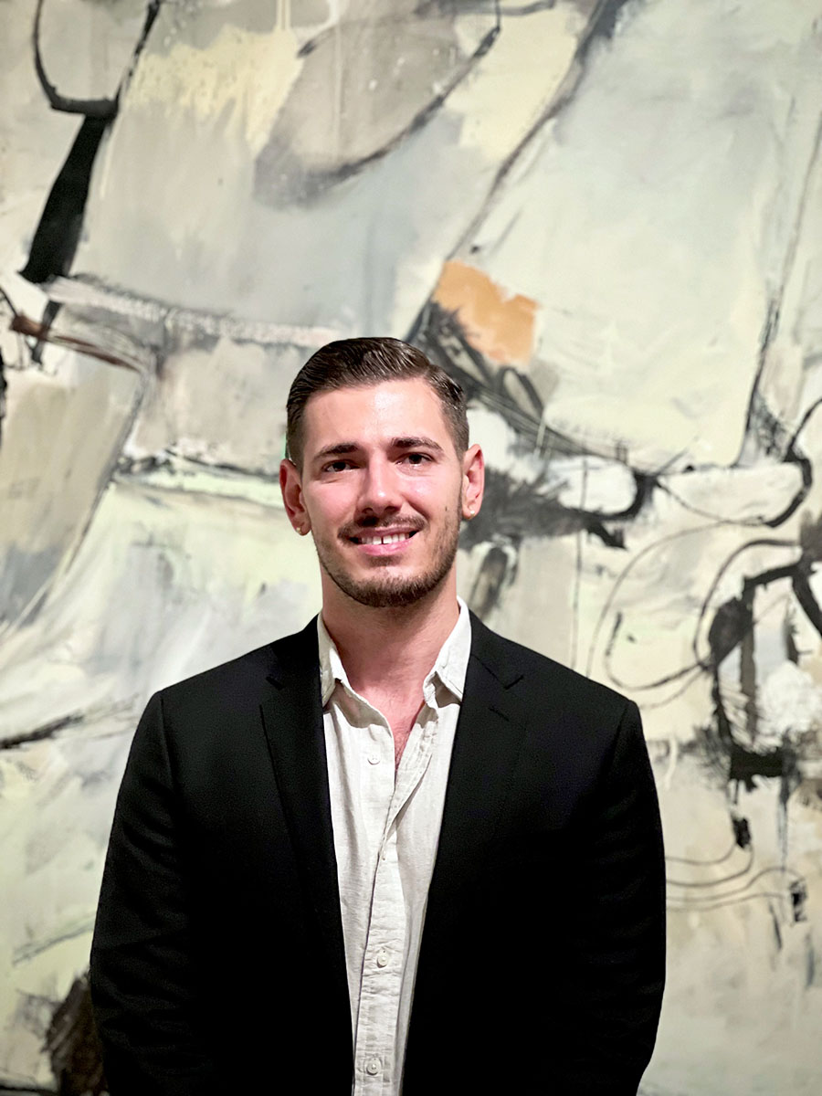 Curator Christian Weguespack, a light skinned man with short dark hair, wearing a dark jacket stands in front of a modern, abstract painting.