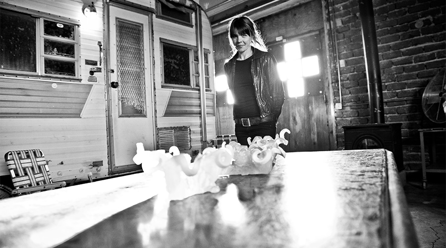 Black and white photograph of the artist standing between a piece of her hand-made glass and one of her Light Ships, a retro recreational vehicle.