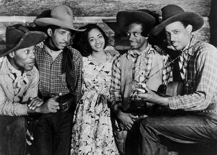 The all-Black cast of Harlem Rides the Range stand close together wearing their costumes.