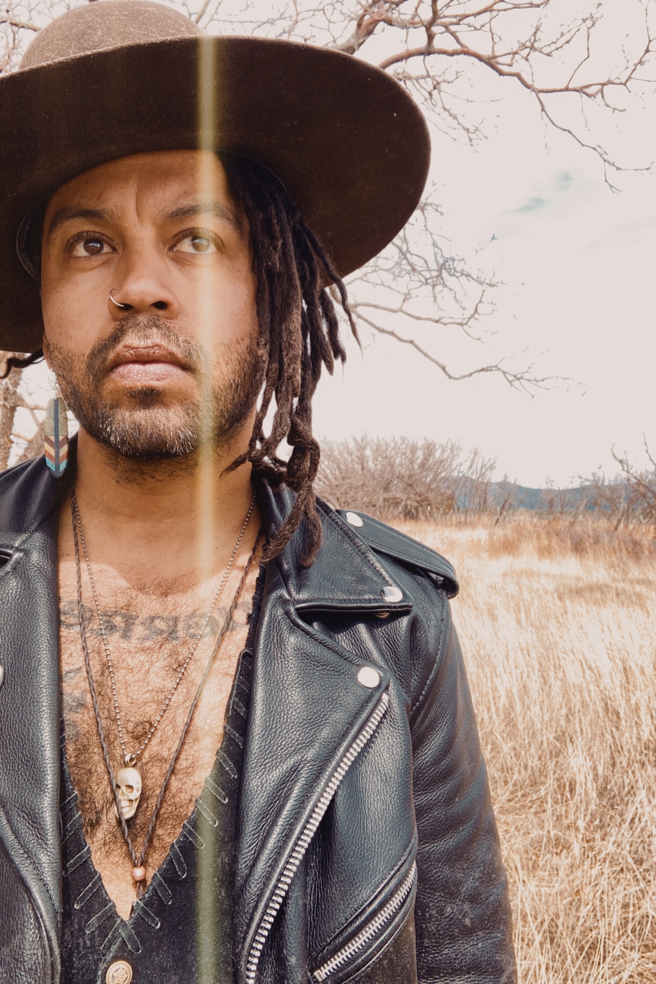 Garry Blackchild, a dark skinned man with dradlocks and stubble, stands in a field wearing a leather jacket and hat. 