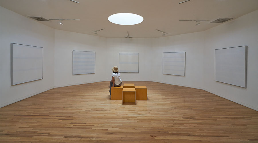 A woman sits on square yellow benchs in the center of an octagonal gallery. She is surrounded by three large, square paintings by Agnes Martin. A round oculus in the ceiling lets in natural light.