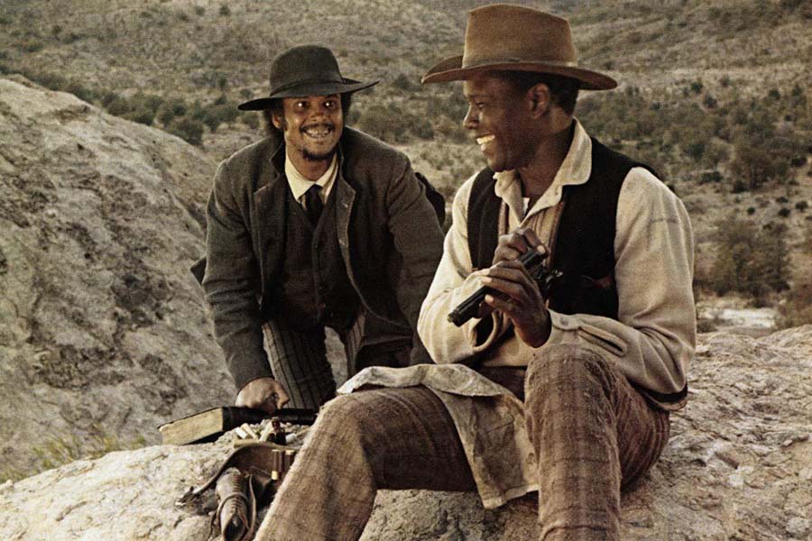 Still from Buck and the Preacher featuring Sidney Poitier and Harry Belafonte wearing cowboy attire and smiling at each other and sitting on a rock.