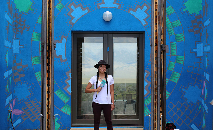 photograph of Lynette Haozous in a black hat holding a paint brush in front of a blue geometric mural